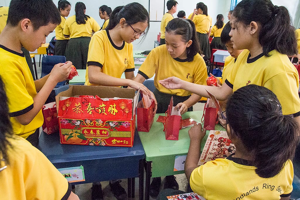 Students in yellow uniforms gather around a table sorting red Chinese New Year gift bags from a decorative box.